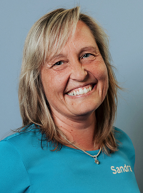 Smiling woman with blonde hair wearing a blue shirt with the name Sandra embroidered on it, against a light blue background.
