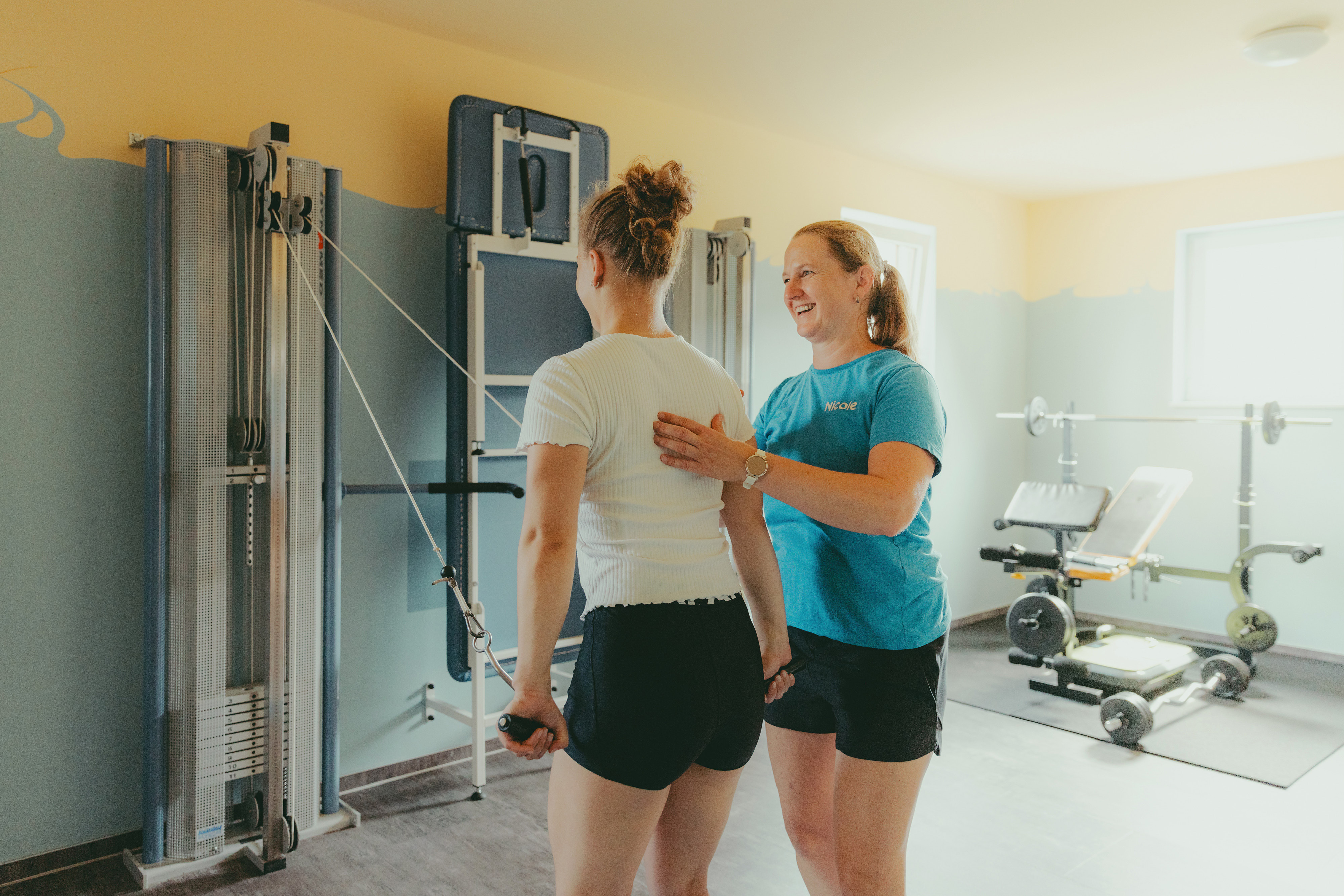 A woman in a blue shirt labeled Nicole coaching another woman in white shirt using cable machine exercise in a gym.