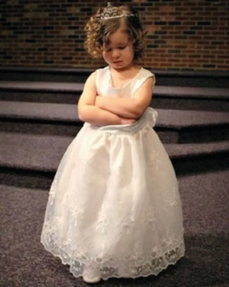 Young girl in a white floral lace dress and tiara standing with her arms crossed, looking down.