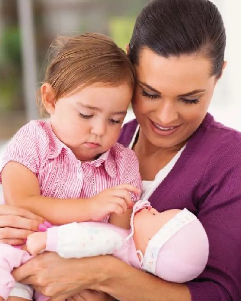 A woman holding a baby doll while a young girl touches the doll’s pacifier.
