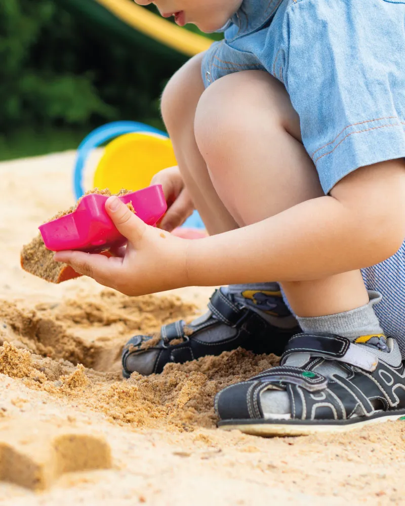 Child wearing black sandals playing with sand using a pink sandbox toy and colorful buckets.
