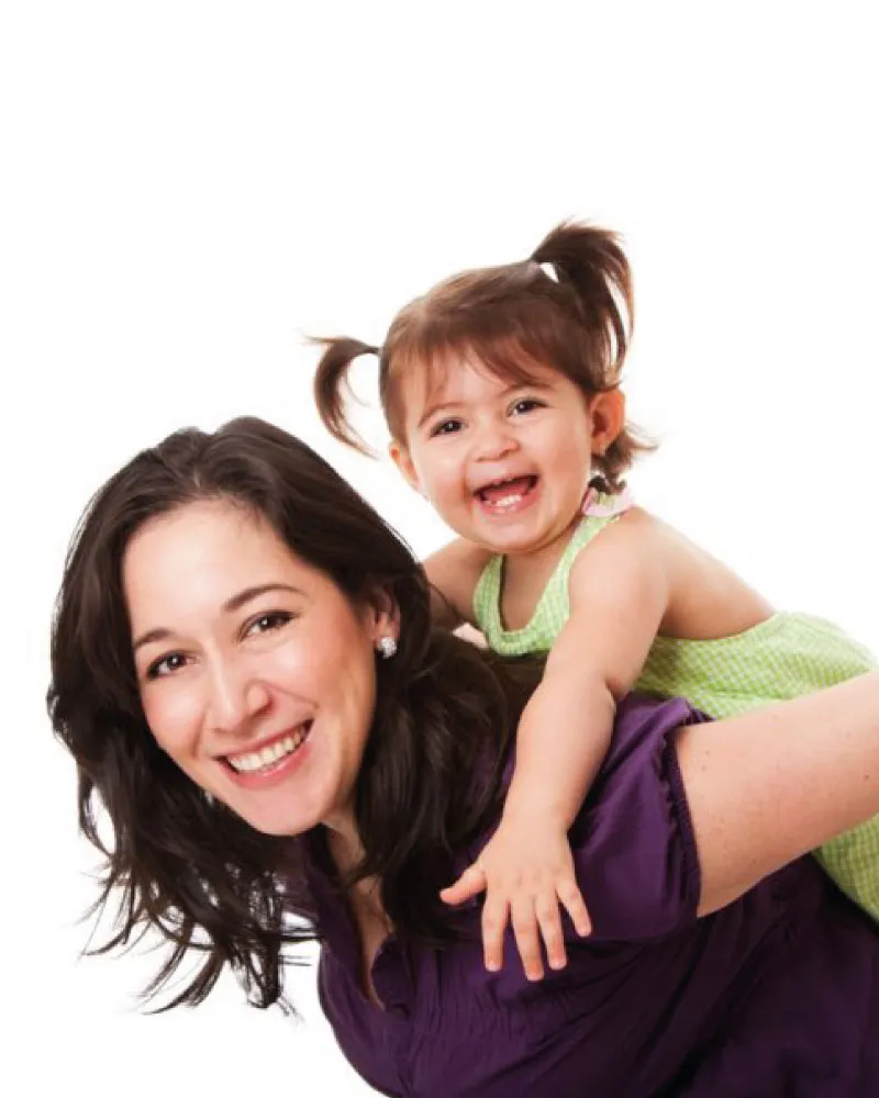 Smiling woman giving a piggyback ride to a happy toddler with pigtails wearing a green dress.