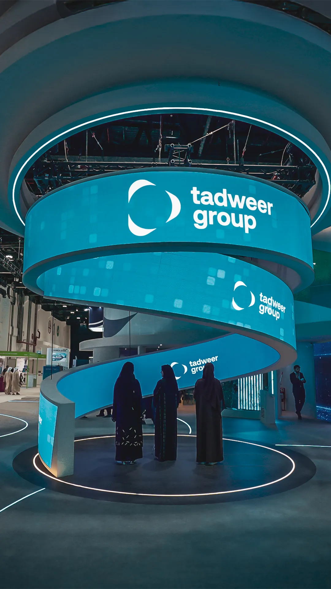 WFES - EcoWaste Three women in traditional black abayas standing inside a circular area beneath a large spiral digital display with 'tadweer group' logo.