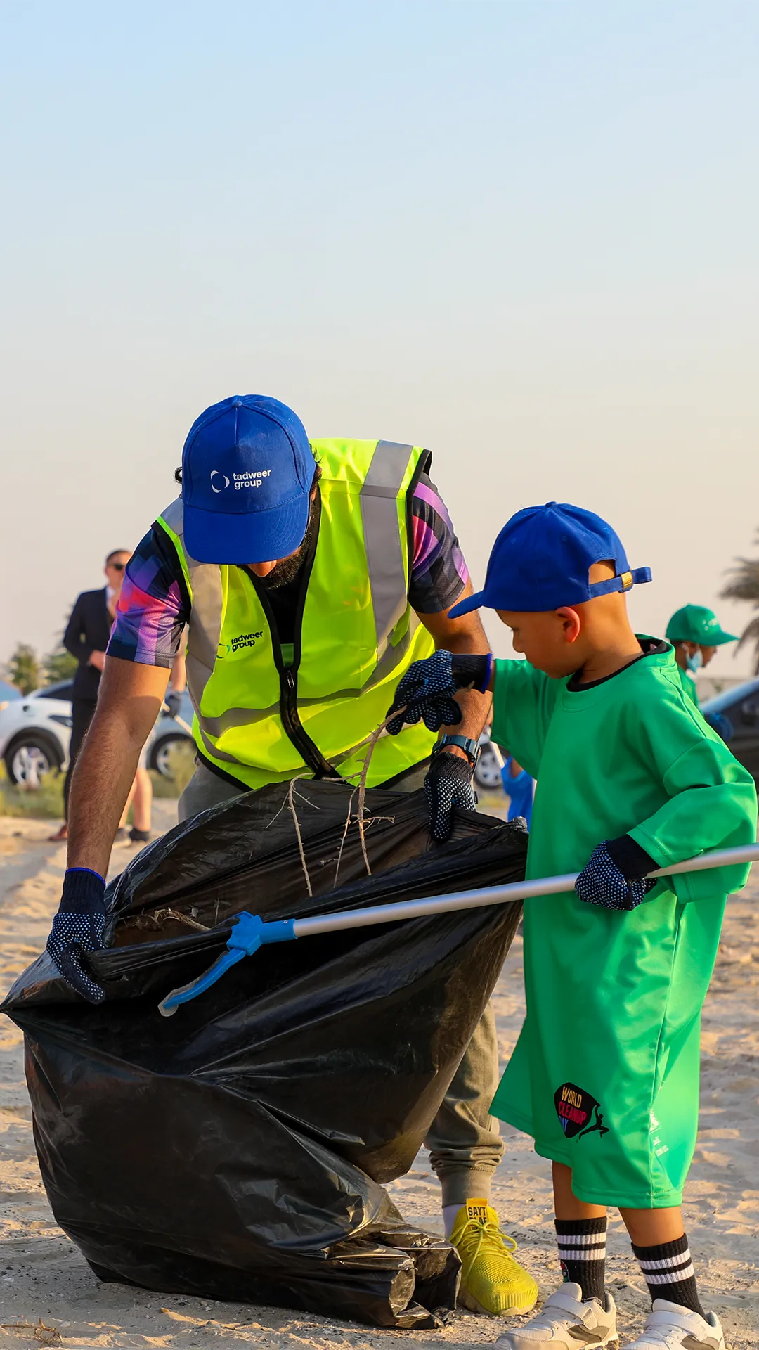 UAE Cleanup campaign by Tadweer Group - Adult and child wearing gloves and caps cleaning up trash on a sandy area using a grabber and a large black garbage bag.