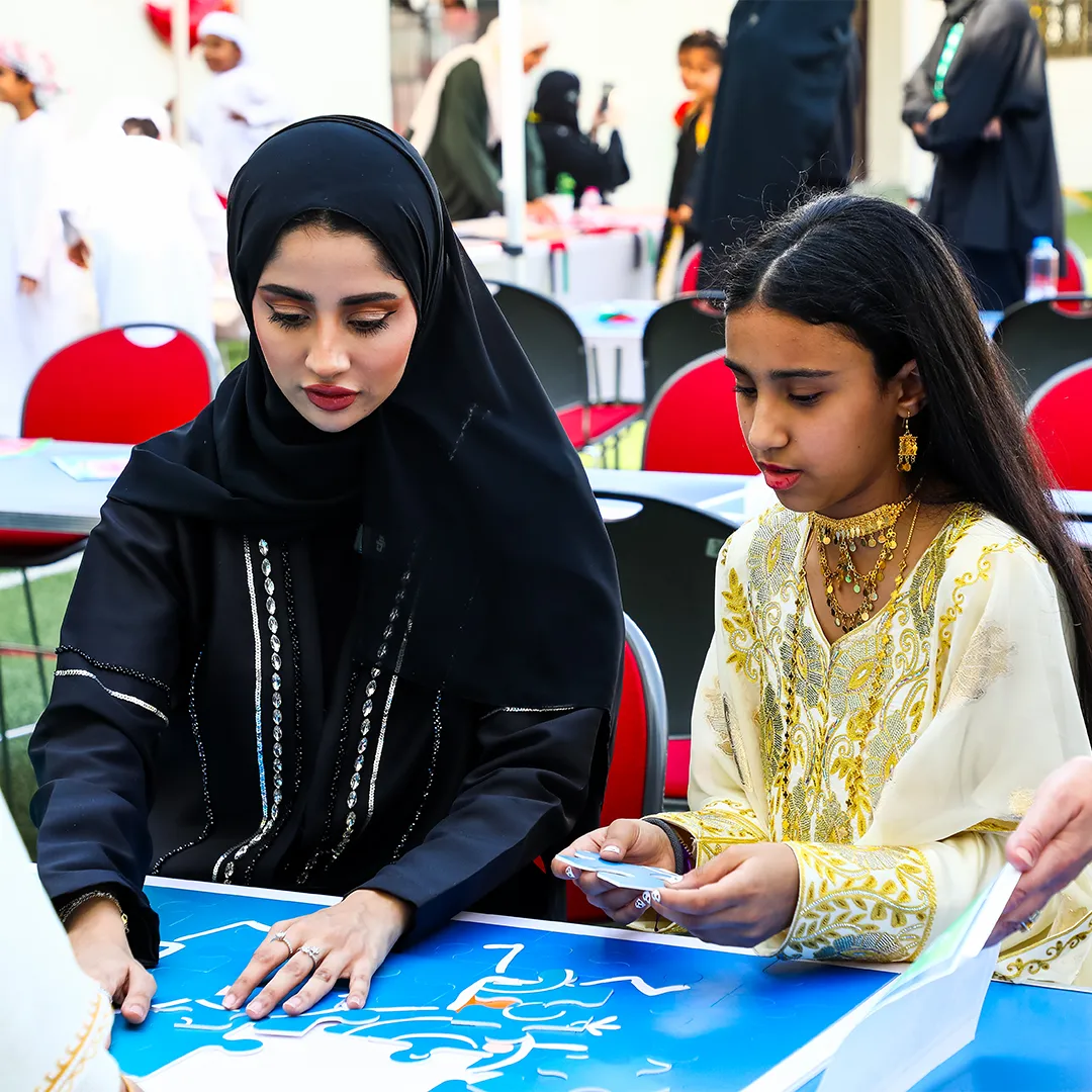 Tadweer Group UAE National day campaign - Two females, one in black abaya and hijab and the other in a gold and white traditional dress, assembling a large blue jigsaw puzzle at an outdoor event.