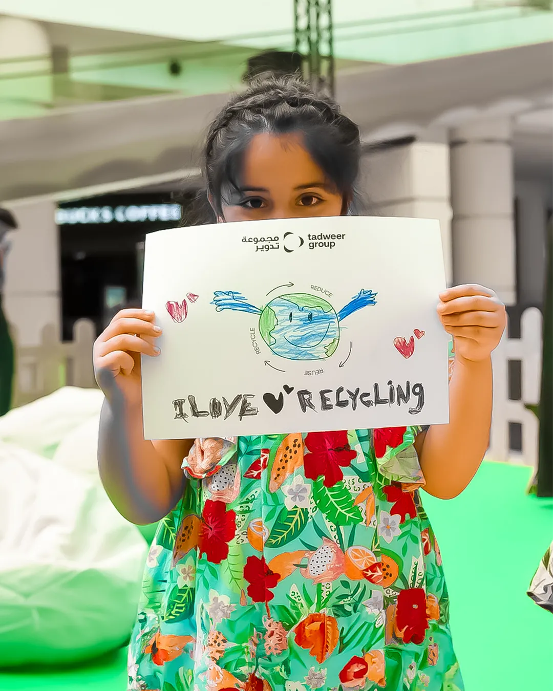 Young girl holding a drawing of a smiling Earth with arms and hearts, text reads 'I love recycling'.