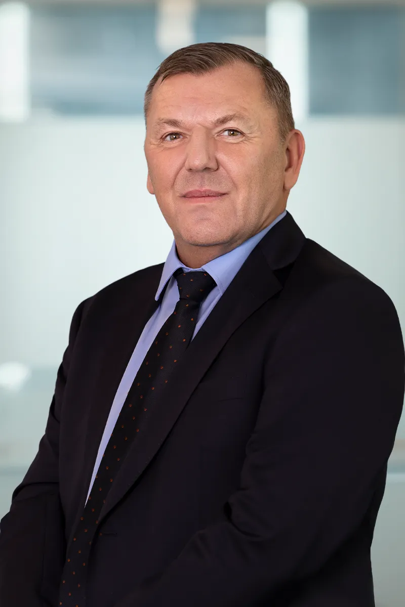Middle-aged man in a dark suit with a blue shirt and patterned tie posing in front of a blurred office background.