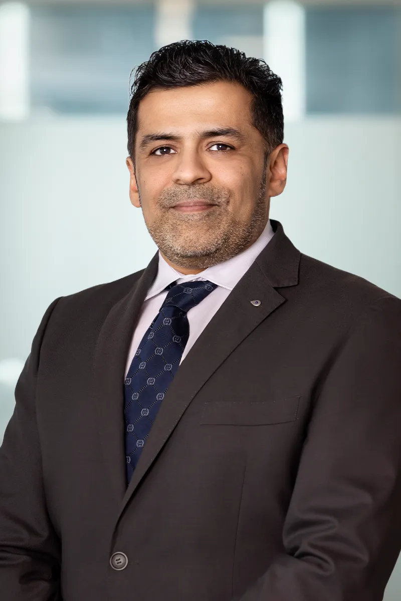 Man in dark suit and patterned navy tie smiling against a blurred indoor background.