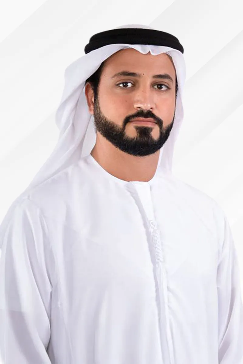 Man with a beard wearing a traditional white kandura and black agal on his head, standing against a light background.