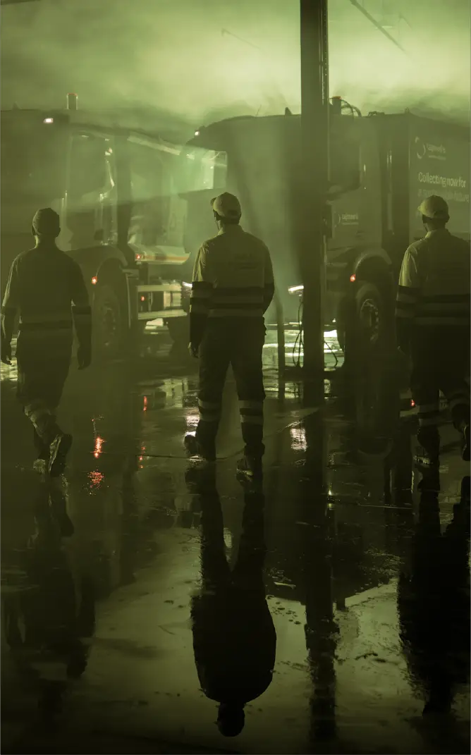 Workers in reflective safety clothing walking near garbage trucks in a foggy, wet industrial area at night.