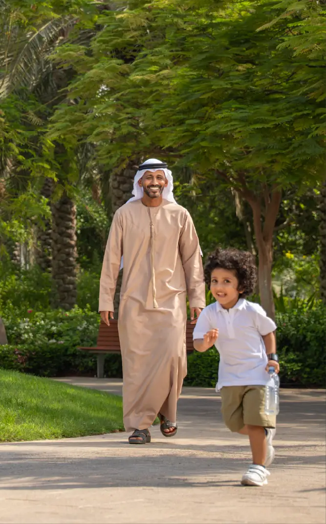 Smiling man in traditional Middle Eastern attire walking behind a young boy running with a water bottle in a lush park.