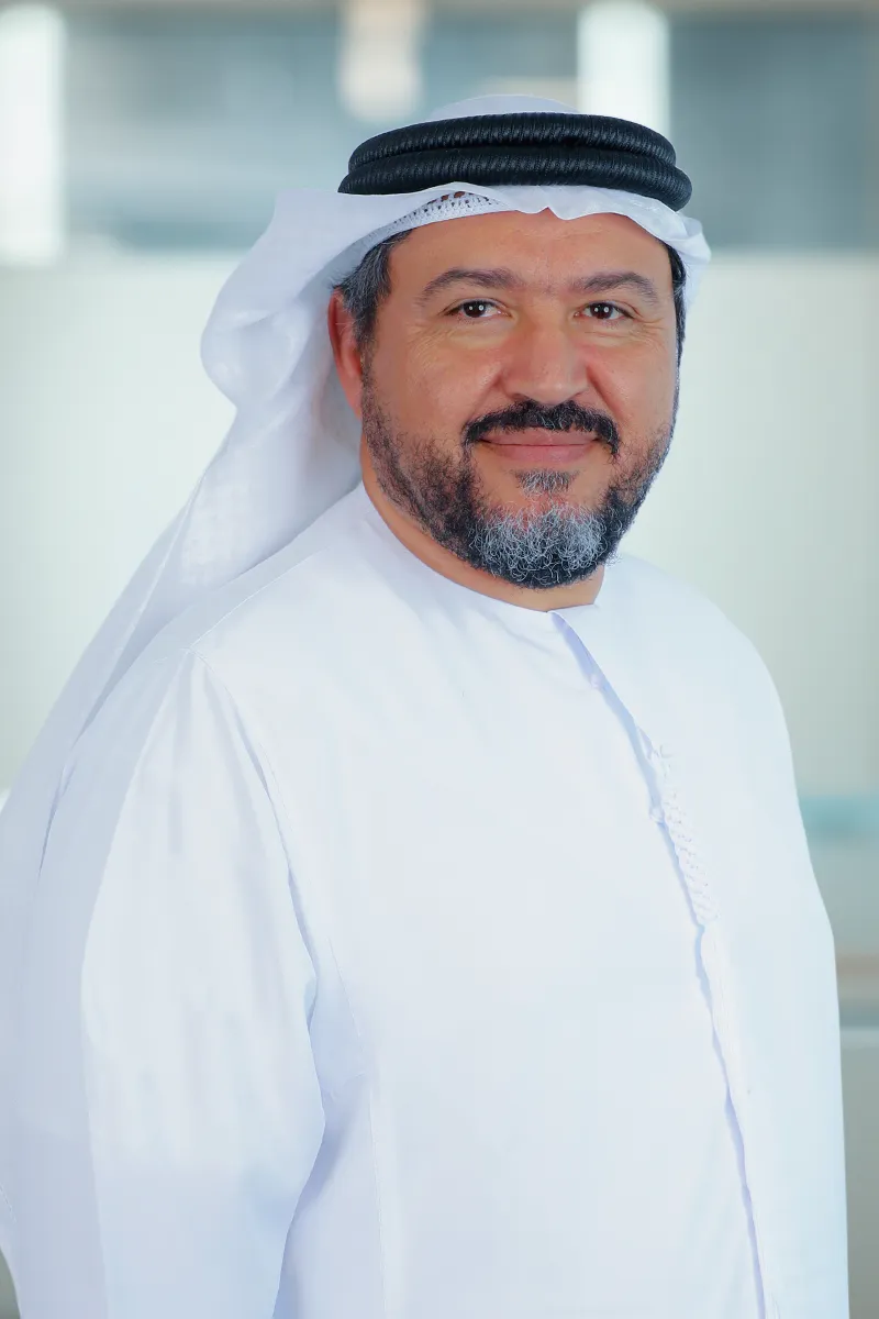 Middle-aged man wearing traditional white Emirati kandura and black agal on his head, smiling softly.