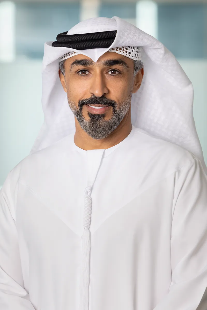 Middle-aged man with salt-and-pepper beard wearing a white kandura and traditional headscarf, smiling softly.