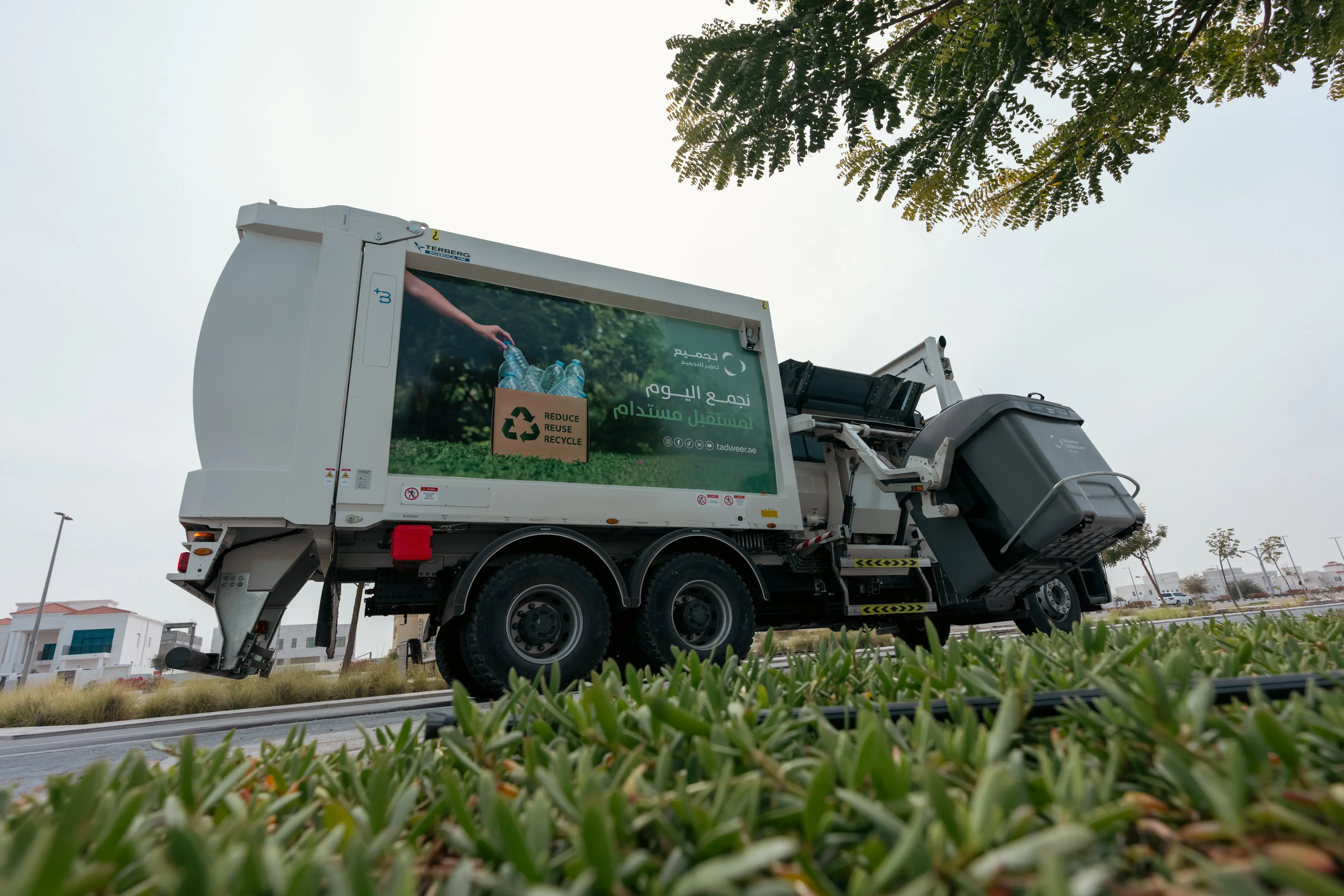 Tajmee'e recycling truck lifting a gray bin near green bushes with a recycling-themed advertisement on its side.