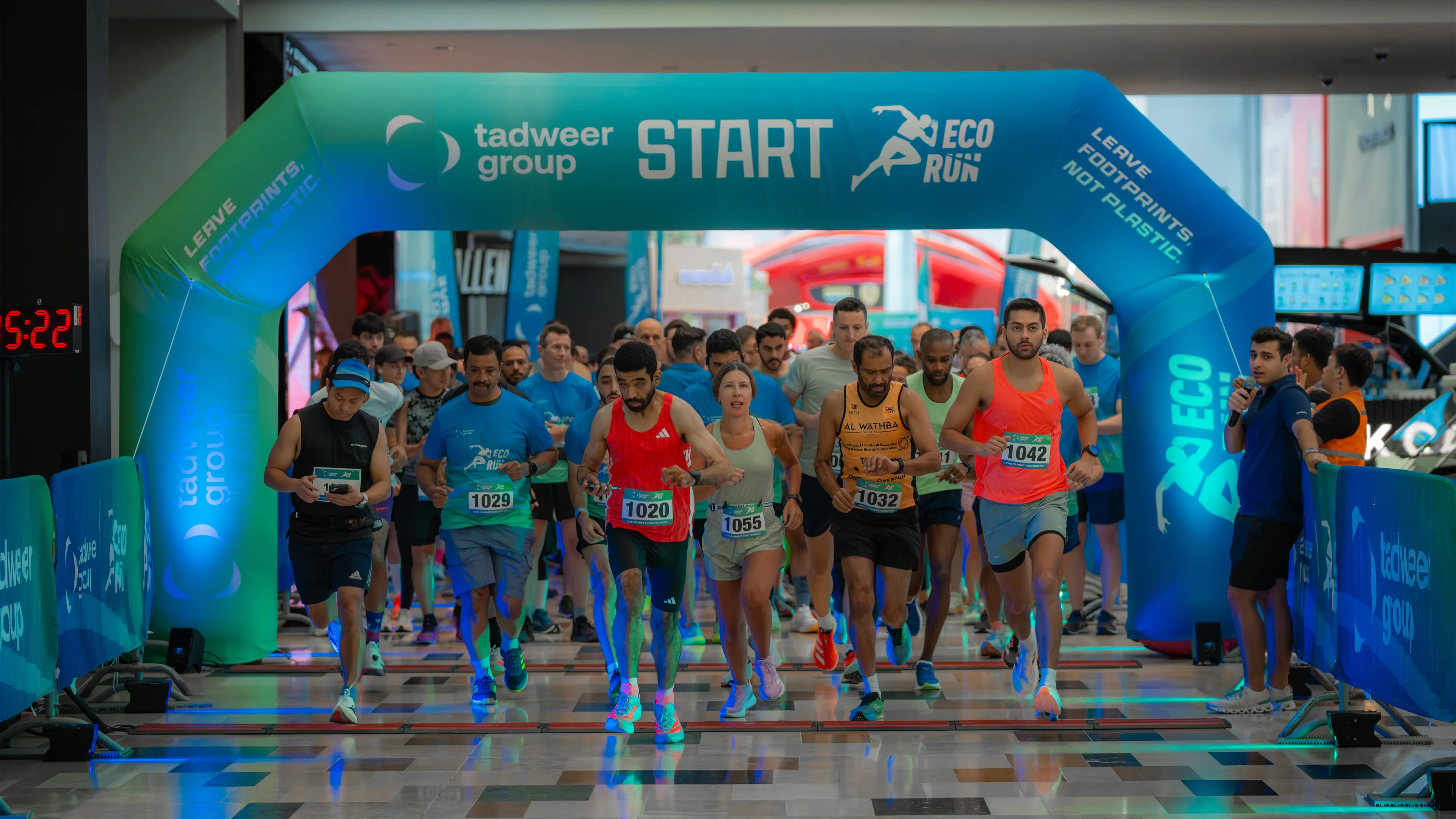 Runners starting an indoor race under a blue arch with 'tadweer group START ECO RUN' text.