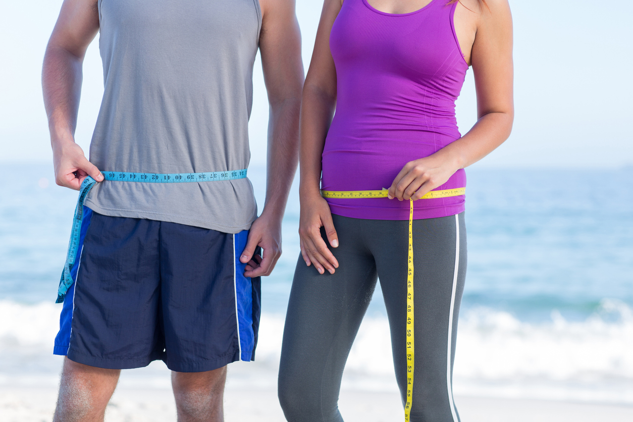 Two adults measuring their waistlines with tape measures outdoors, symbolizing the start of a weight loss journey.