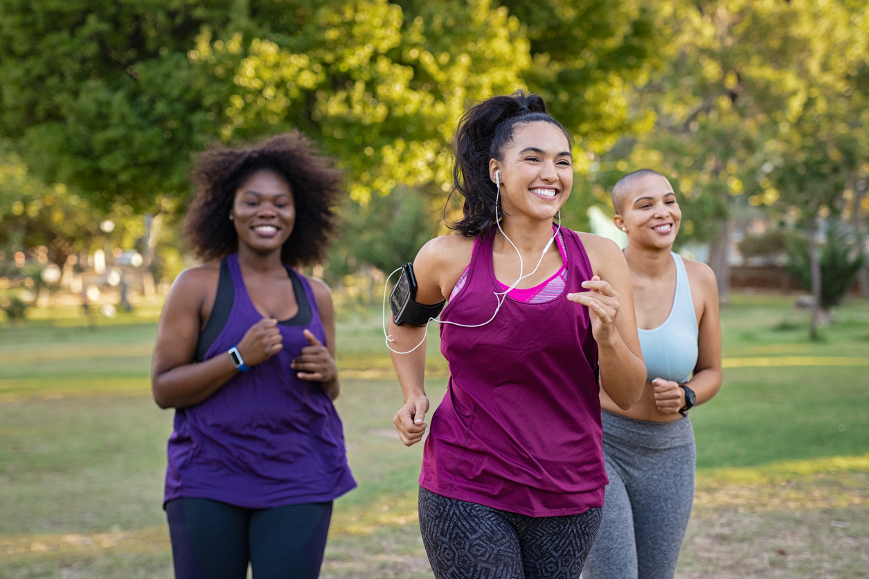 Three women jogging together in a sunny park, smiling in fitness wear and representing active, healthy living.
