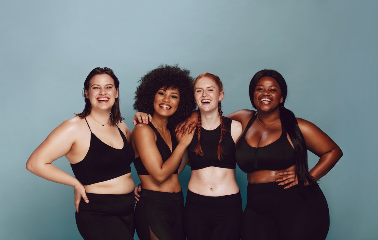 Four women in black sportswear smiling with arms around each other, celebrating fitness and body positivity.