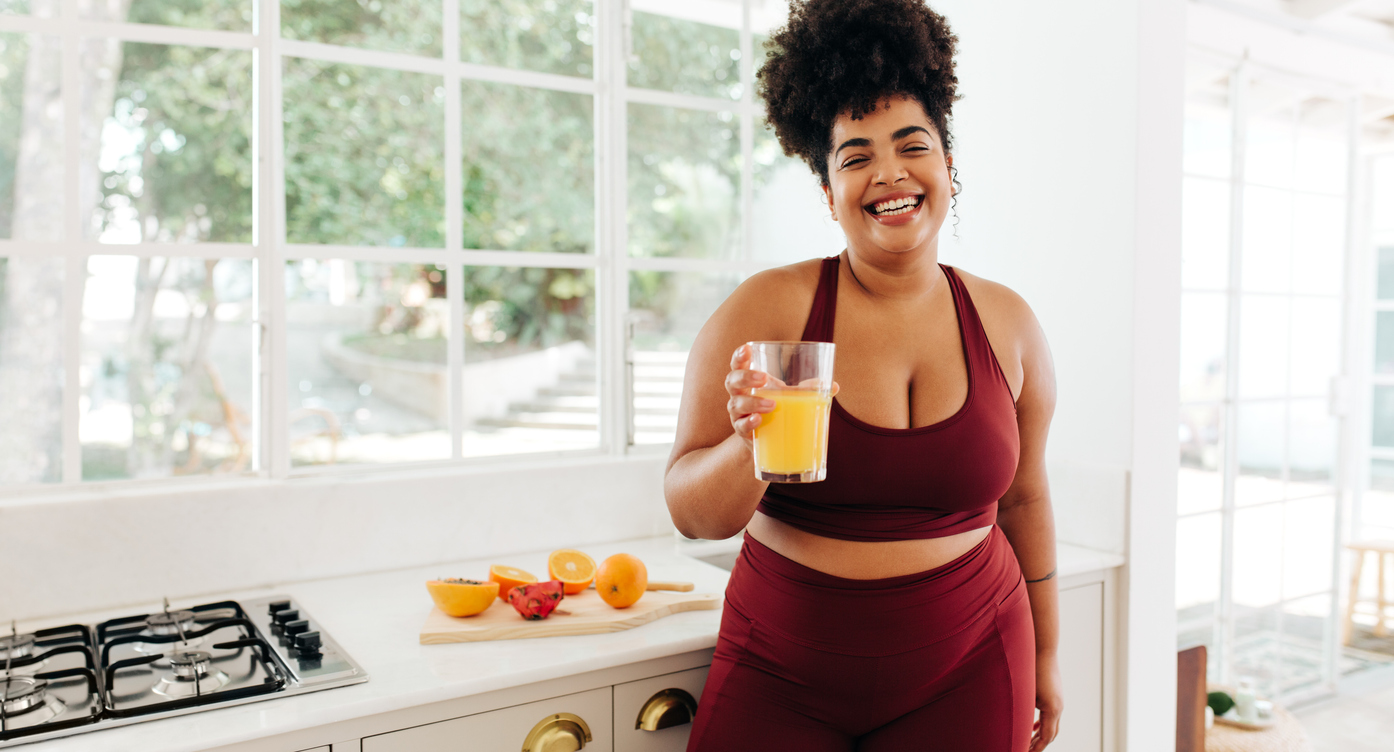 Smiling woman in red activewear holding a glass of fresh orange juice in a bright kitchen, representing daily wellness and energy.