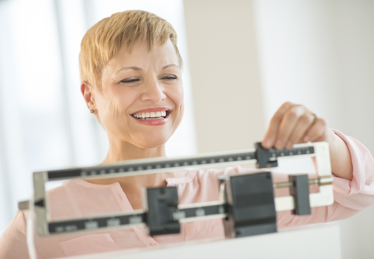 Middle-aged woman smiling while standing on a bathroom scale, representing monitored weight loss progress through provider-guided care.