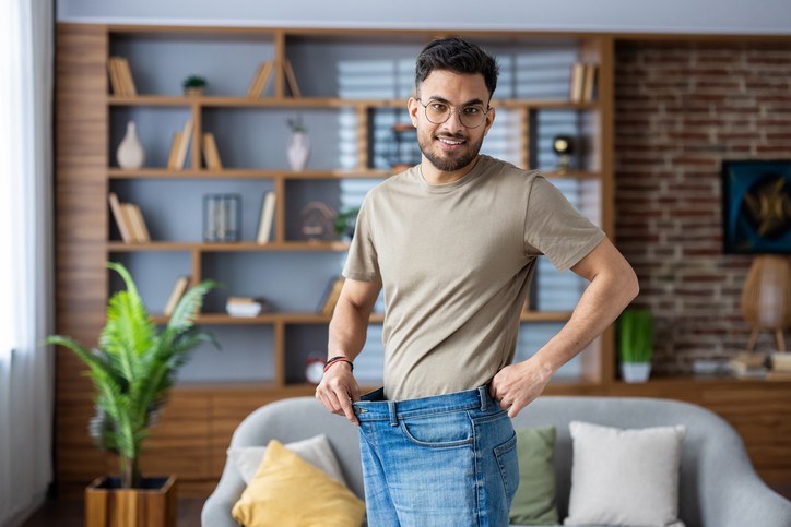 Man at home holding oversized jeans to demonstrate noticeable weight loss and body transformation progress.