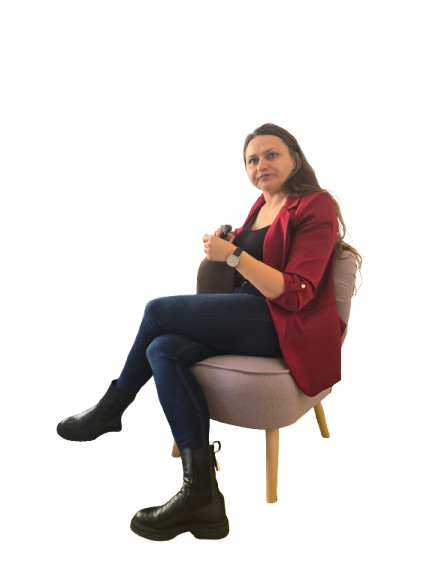 Woman wearing a red blazer, black top, jeans, and black boots sitting on a light pink chair with legs crossed.