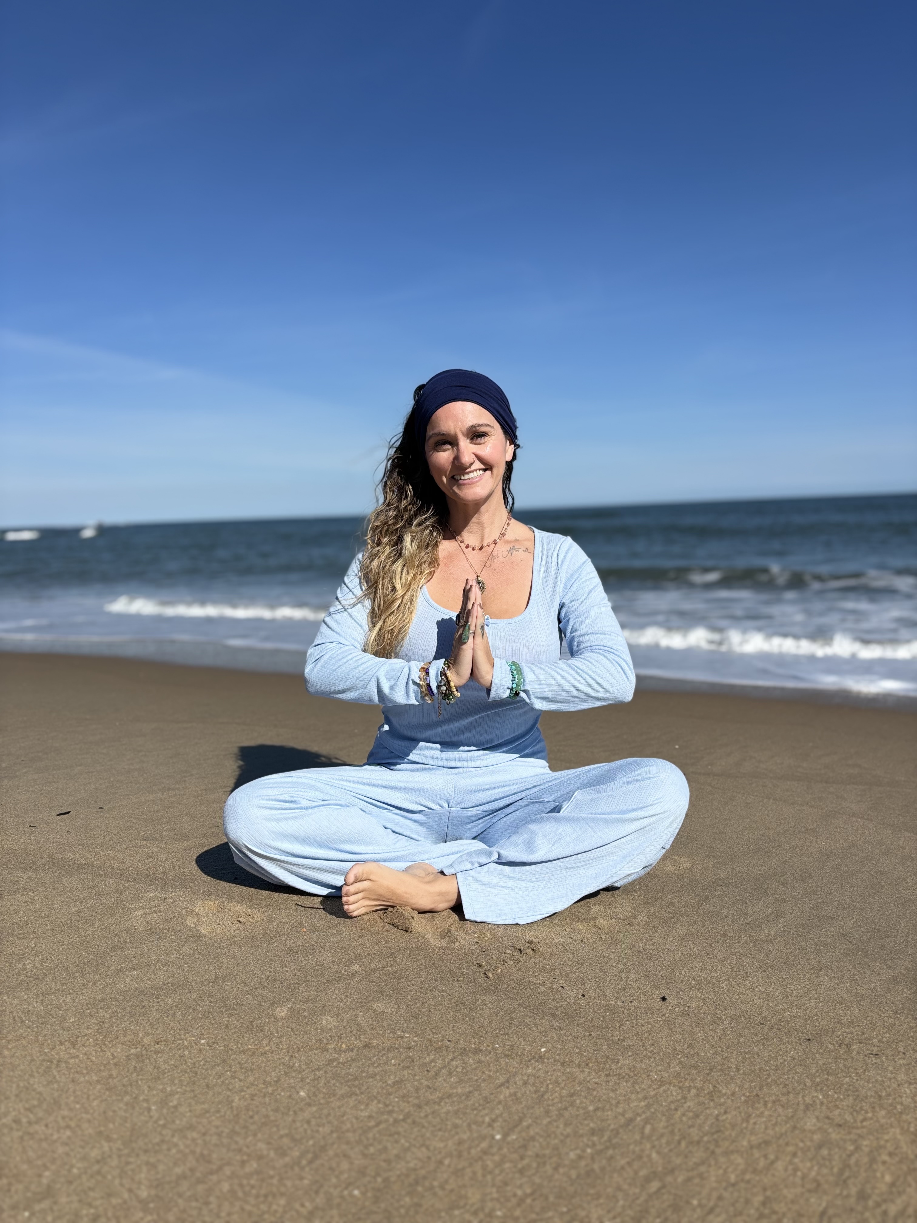 Smiling woman in light blue outfit and headband sitting cross-legged on sandy beach with hands in prayer pose.