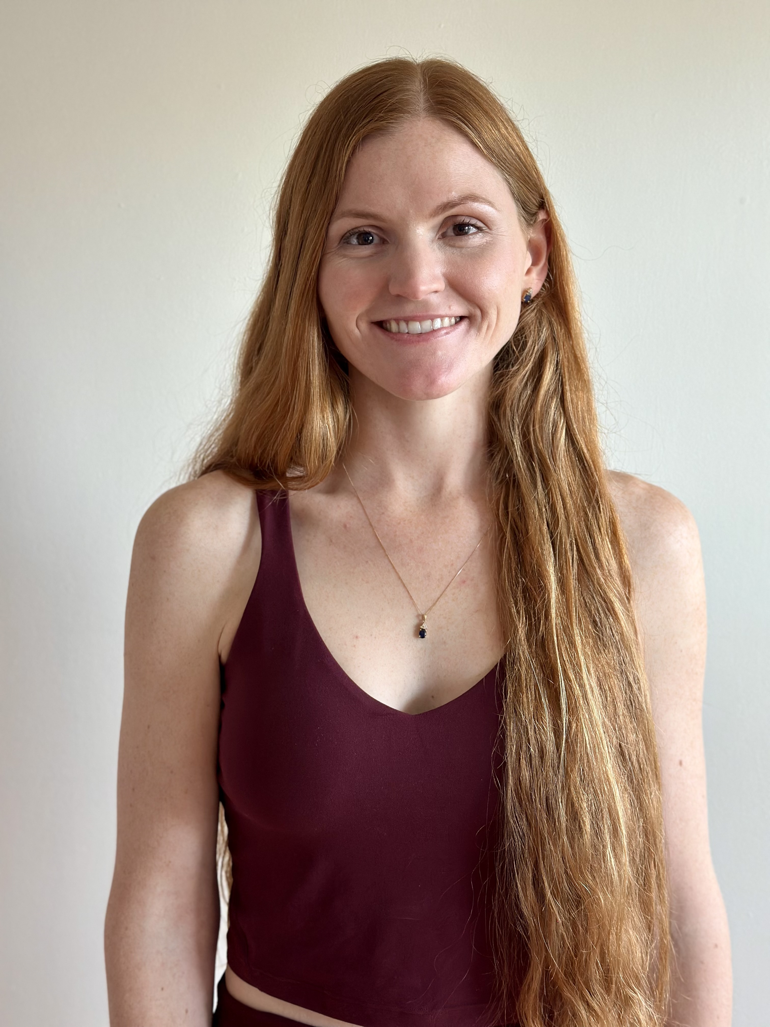 Smiling woman with long red hair wearing a sleeveless burgundy top and a pendant necklace against a plain background.