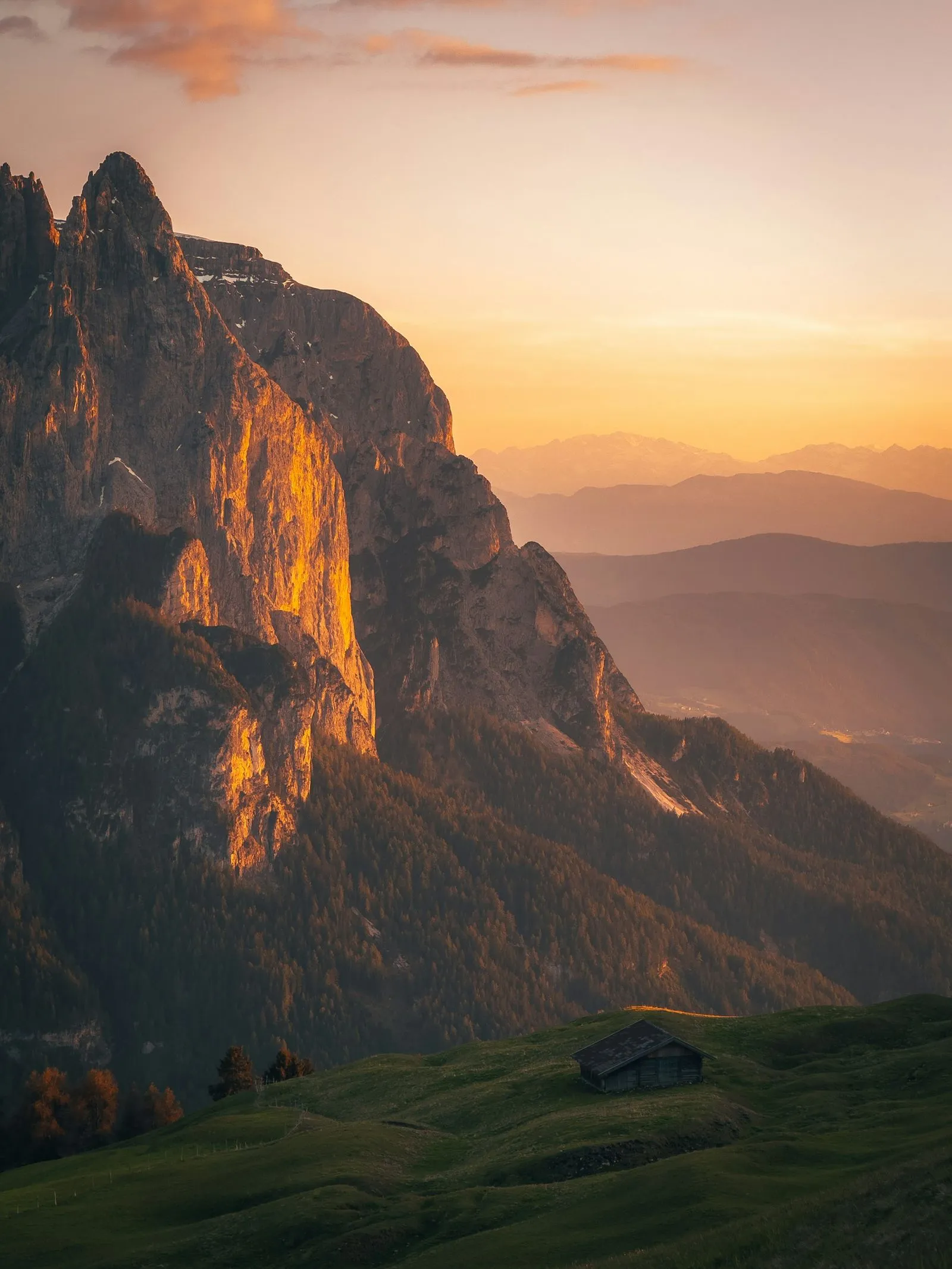 Mountain cliff illuminated by golden sunset with green hills and a solitary cabin in the foreground.