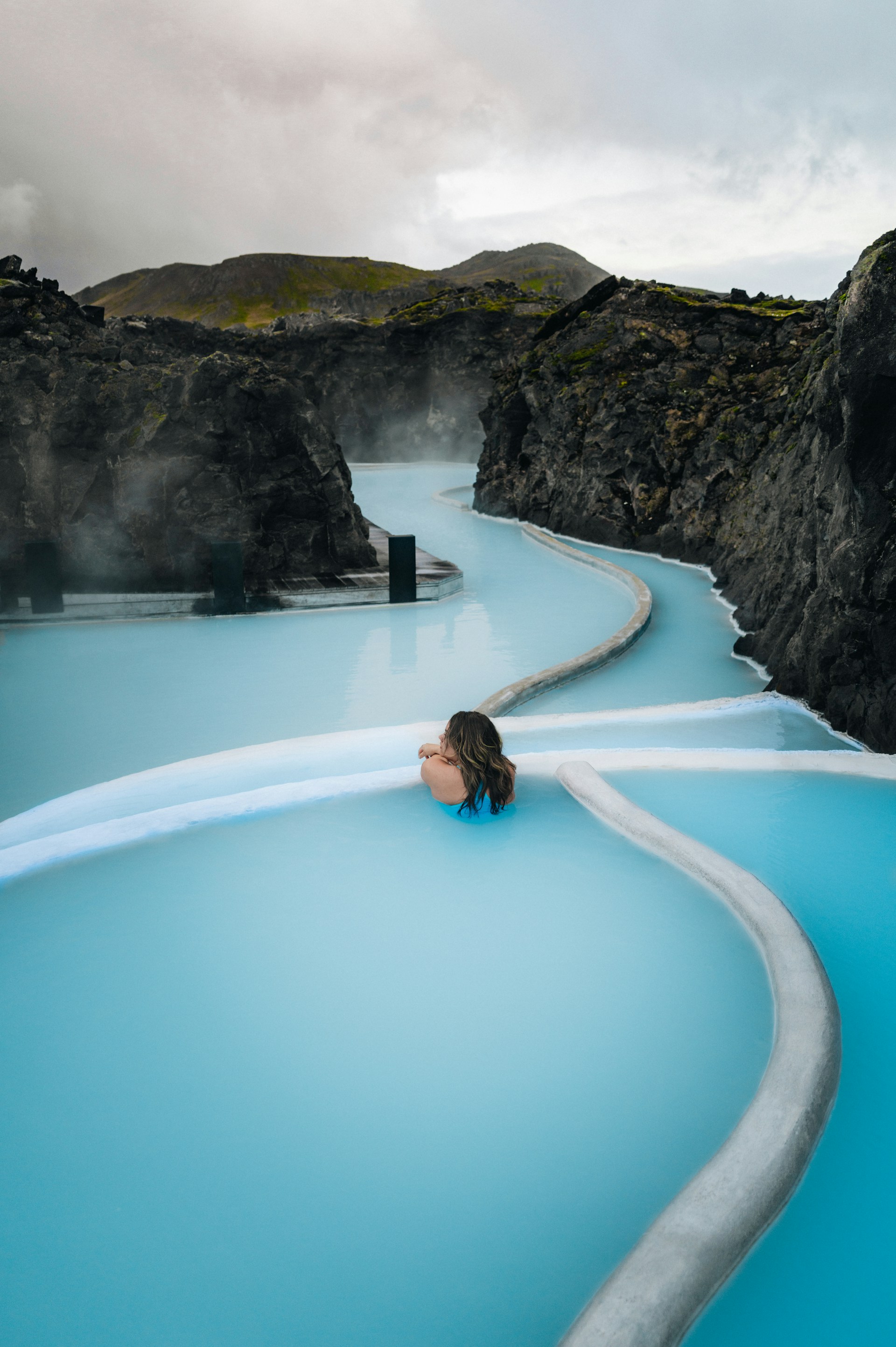Person relaxing in milky blue geothermal pool surrounded by black volcanic rocks under a cloudy sky.