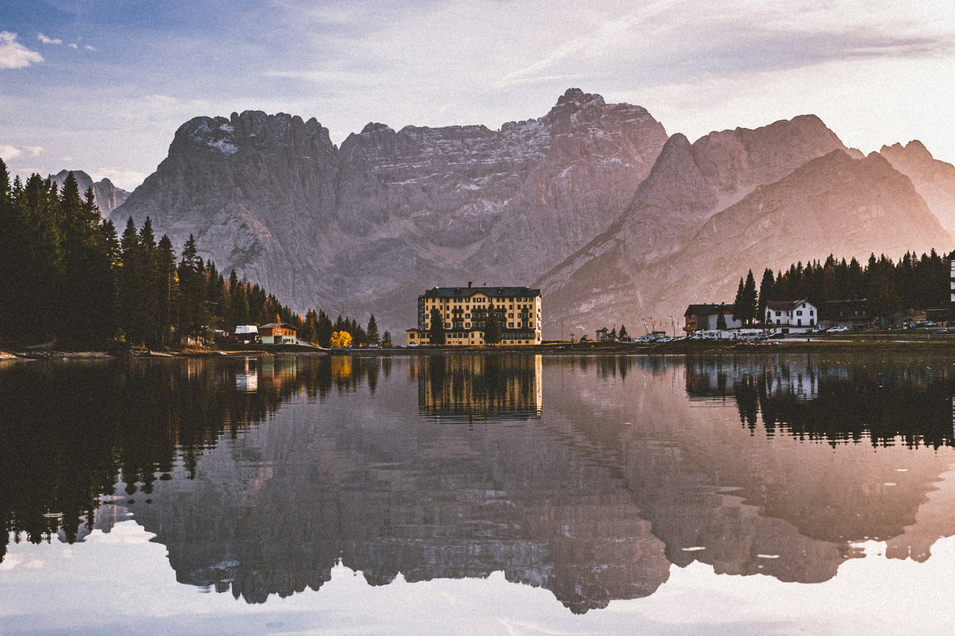 Mountain lake at sunset reflecting pine trees and buildings with large rocky peaks in the background.