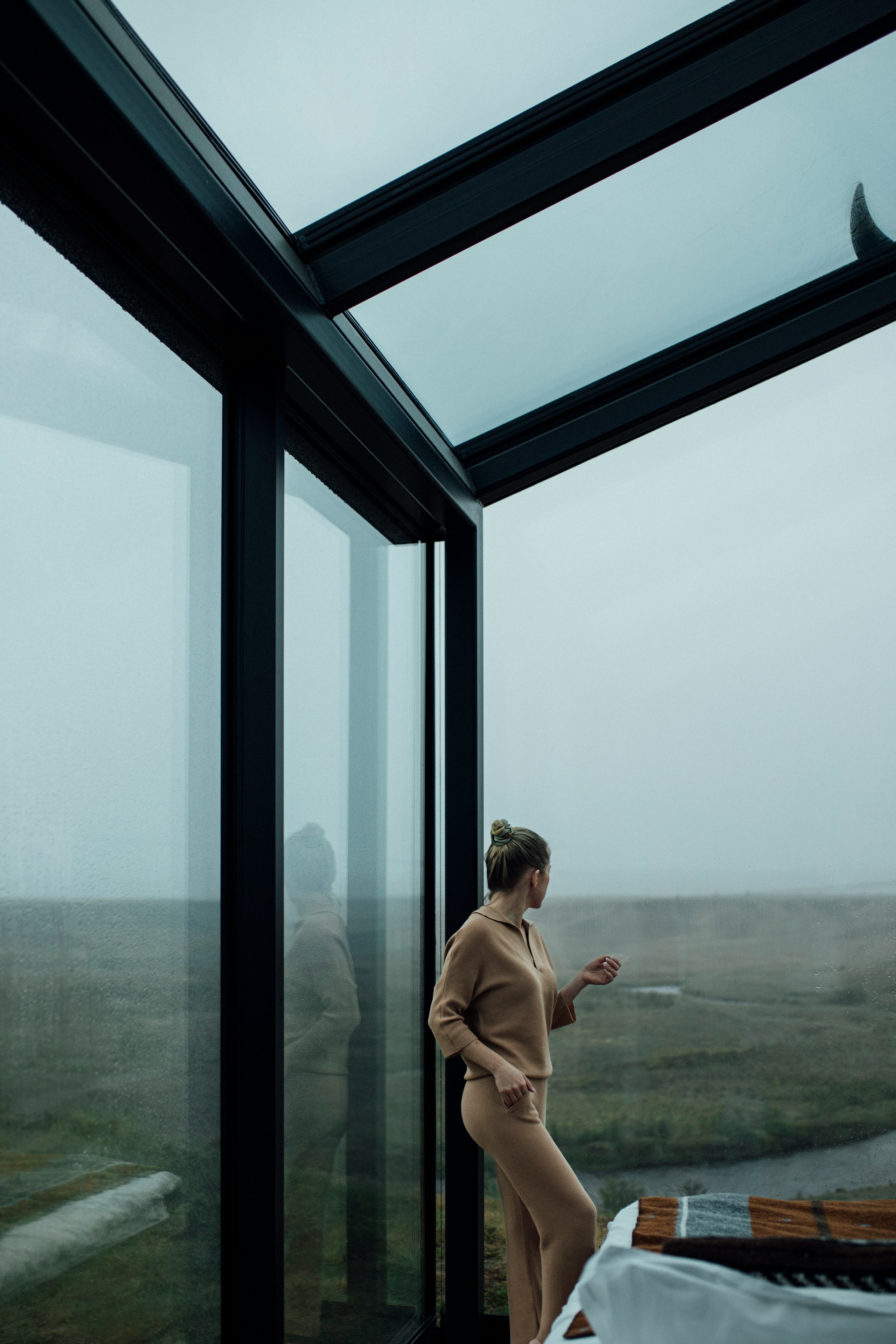 Woman in beige loungewear standing beside large glass windows, looking out at a foggy natural landscape.
