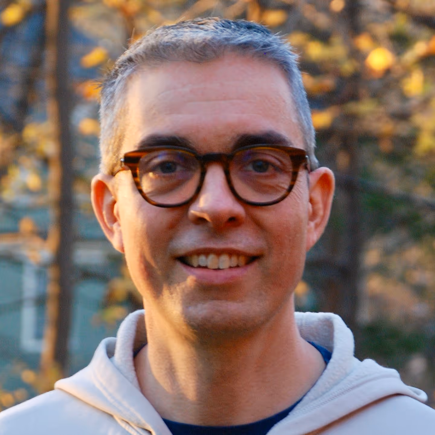 Smiling man wearing tortoiseshell glasses and a light hoodie outdoors with blurred autumn foliage background.