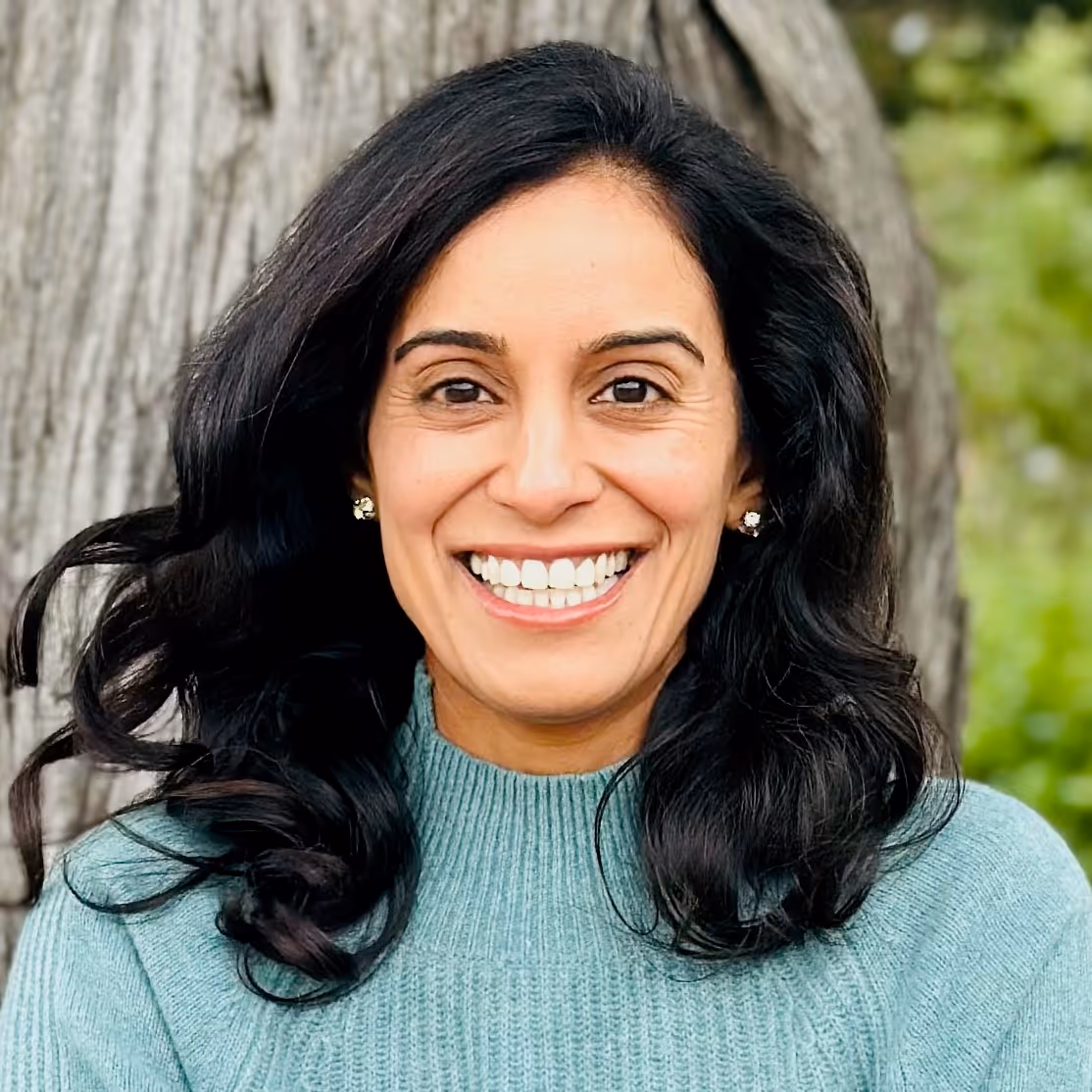 Smiling woman with long dark hair wearing a light green sweater standing outdoors by a tree.