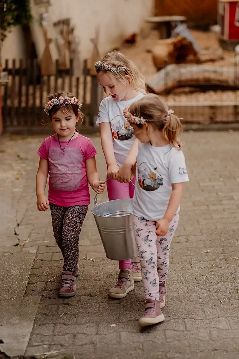 Drei Mädchen mit Blumenkränzen tragen gemeinsam einen Eimer über den Hof in Hannas-Hofglück-T-Shirts