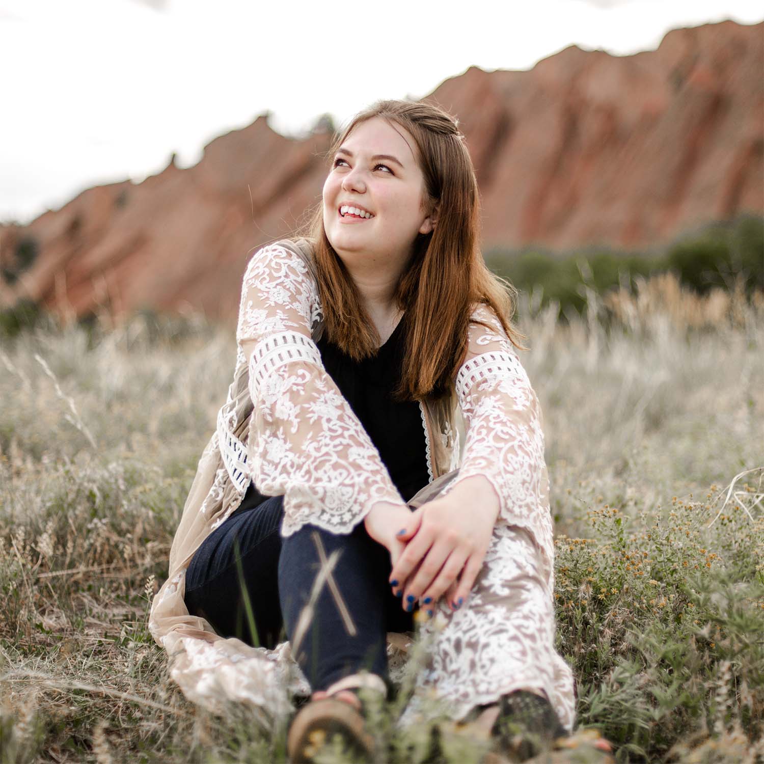 Smiling young woman in a white lace cardigan sitting in a grassy field with red rock formations behind.