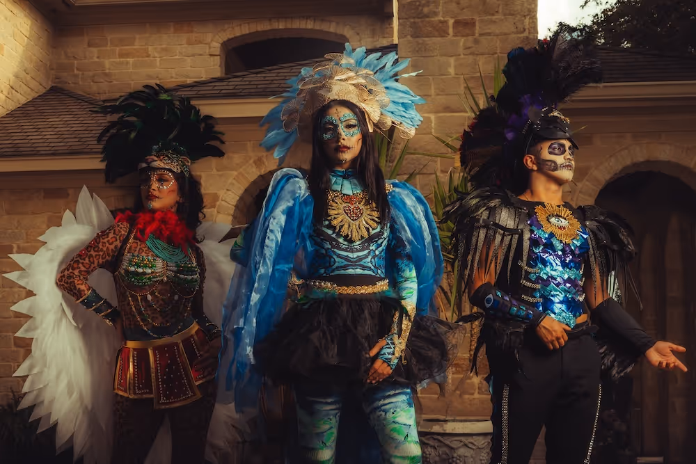 Three people dressed in elaborate Day of the Dead costumes with face paint and feathered headdresses standing in front of a stone building.