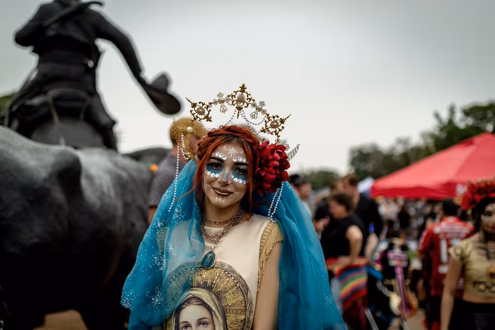 Person dressed in colorful Day of the Dead makeup and costume with a gold headdress, blue veil, and red flower, standing outdoors at an event.