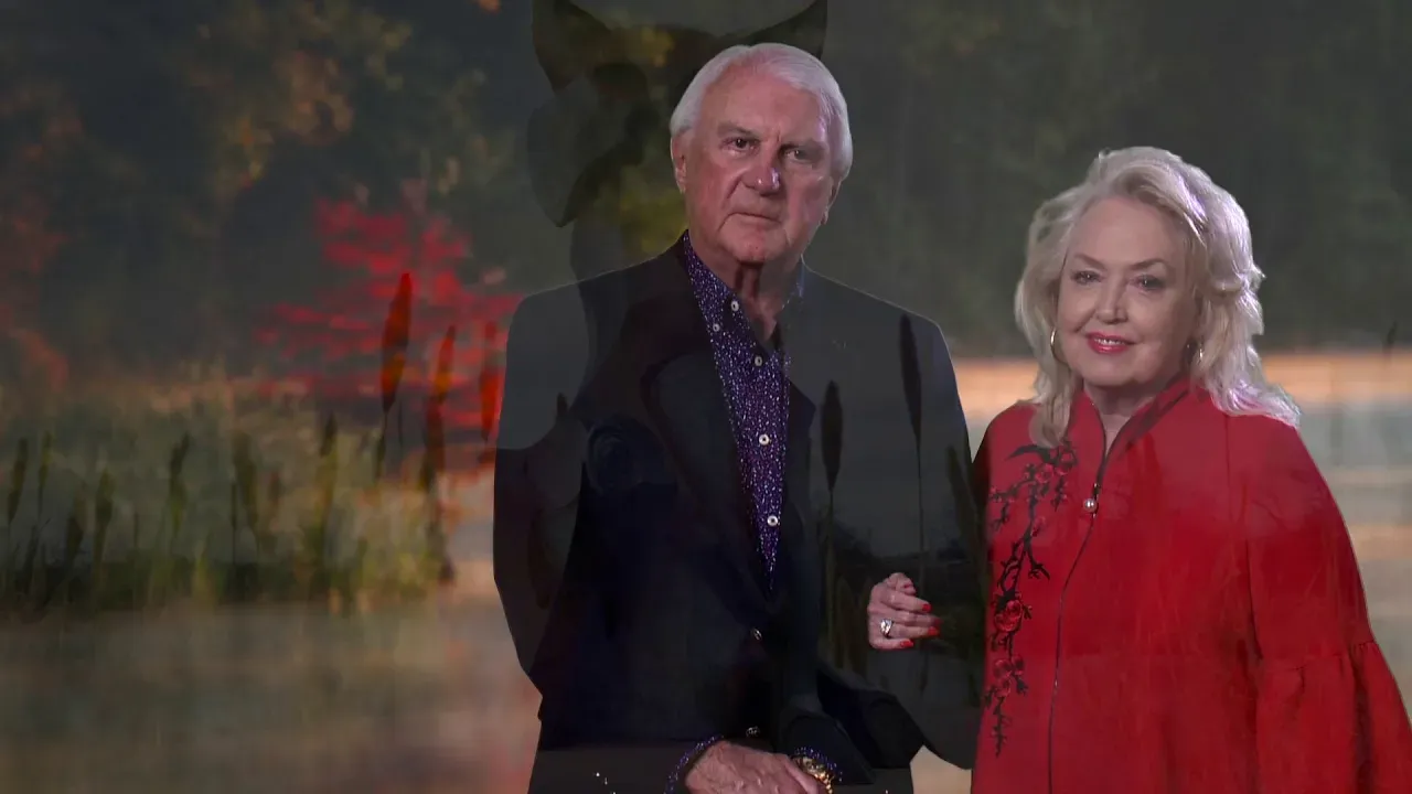 Older man and woman standing outdoors with autumn foliage in the background; the woman wears a red jacket with floral embroidery and smiles.