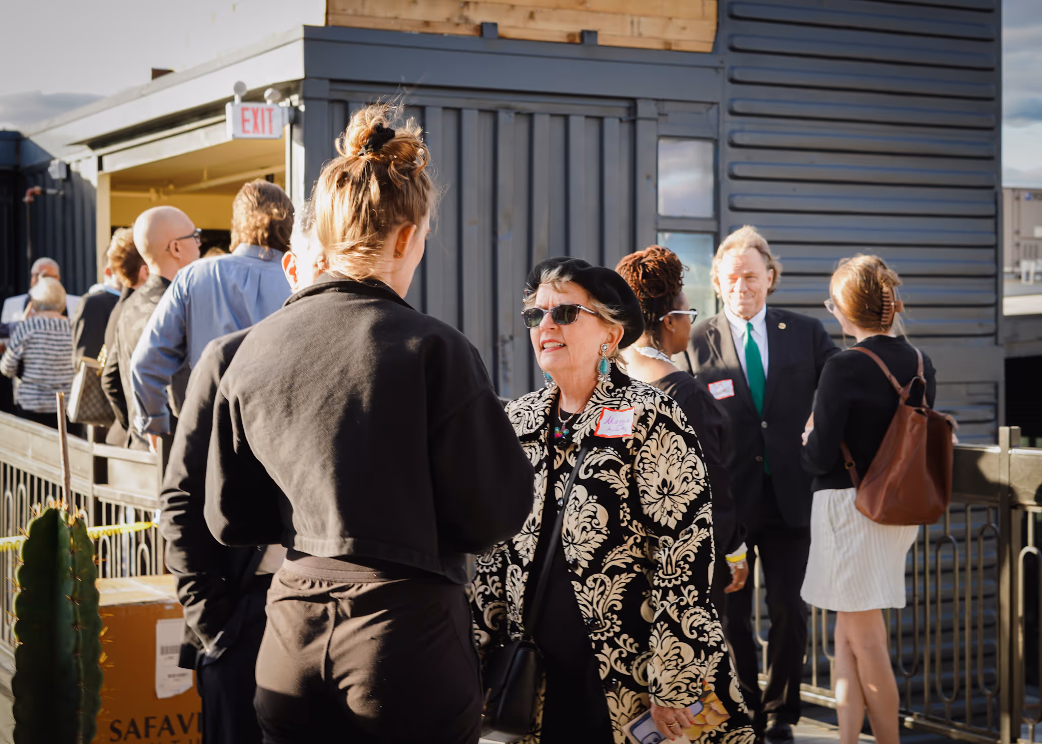 Group of people engaging in conversation outdoors near a gray building entrance with an exit sign.