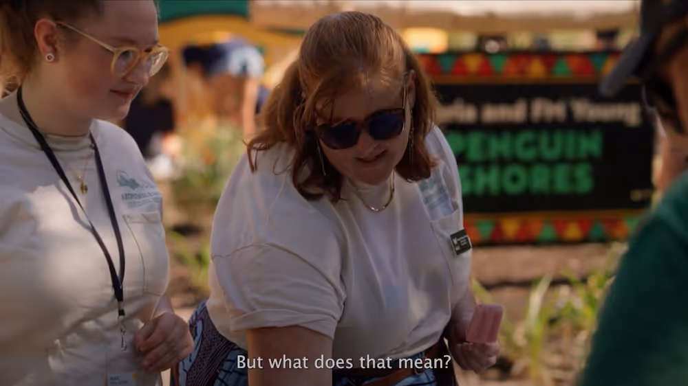 Two women in white shirts talking outdoors near a sign that says Penguin Shores, with one woman wearing sunglasses and subtitles reading 'But what does that mean?'