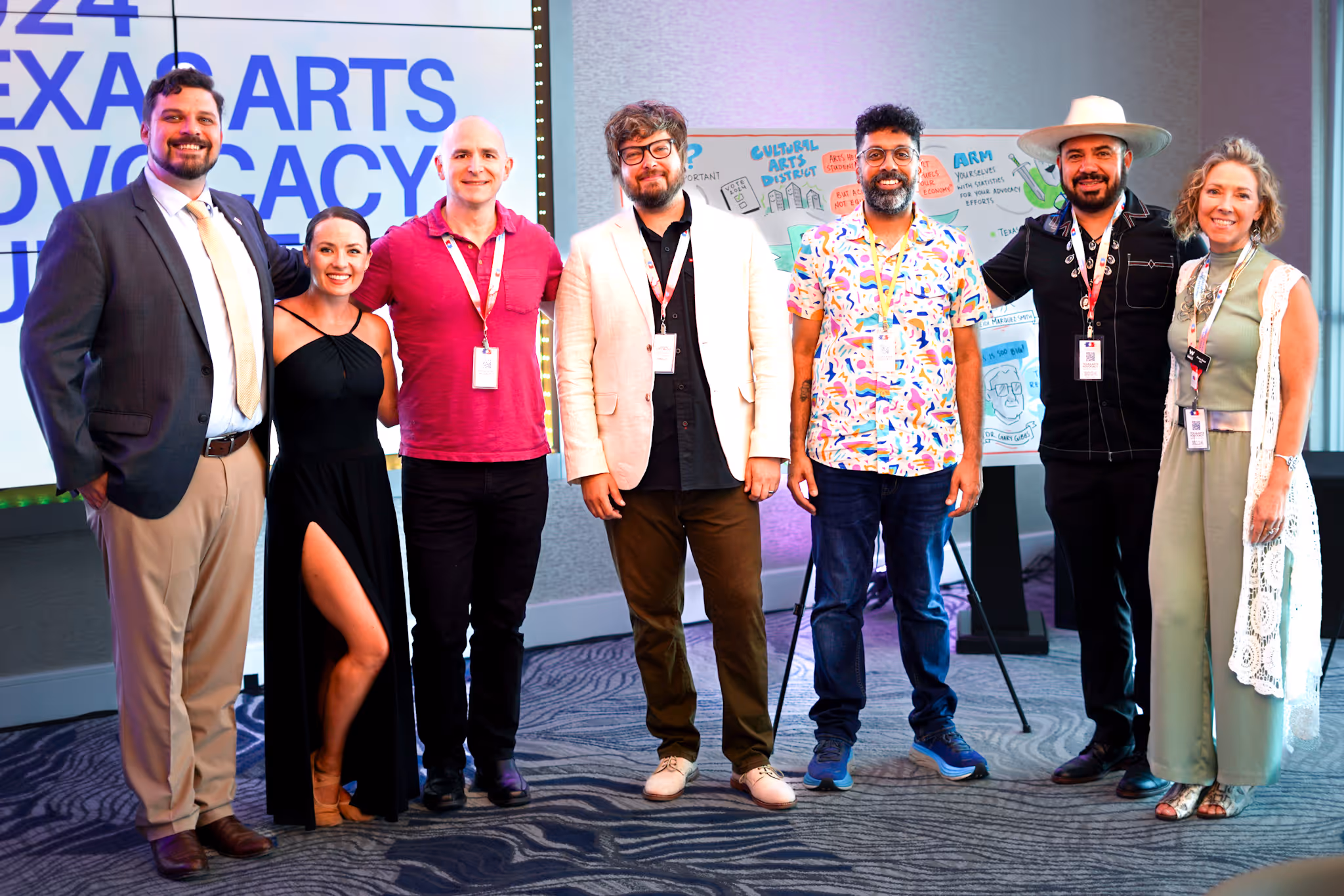 Seven people wearing event badges standing together in a conference room with a presentation board and large screen behind them.