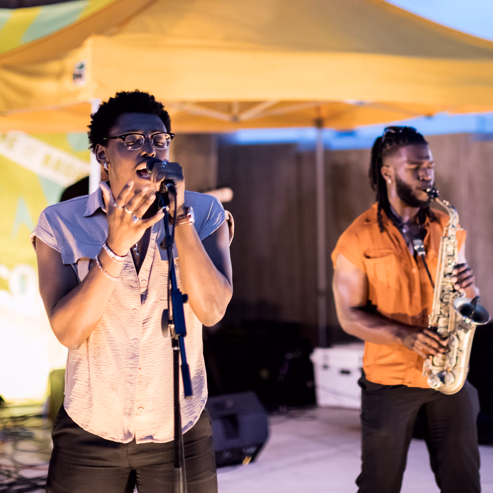 Man singing passionately into a microphone with another man playing the saxophone in the background at an outdoor event under a yellow canopy.