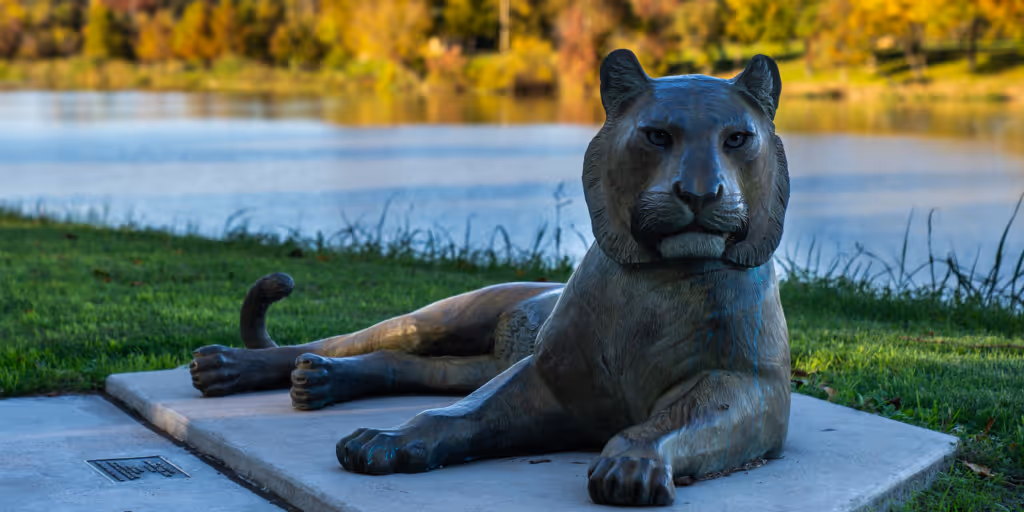 Bronze lion sculpture lying on a stone platform near a lake with grassy shore and trees in the background.