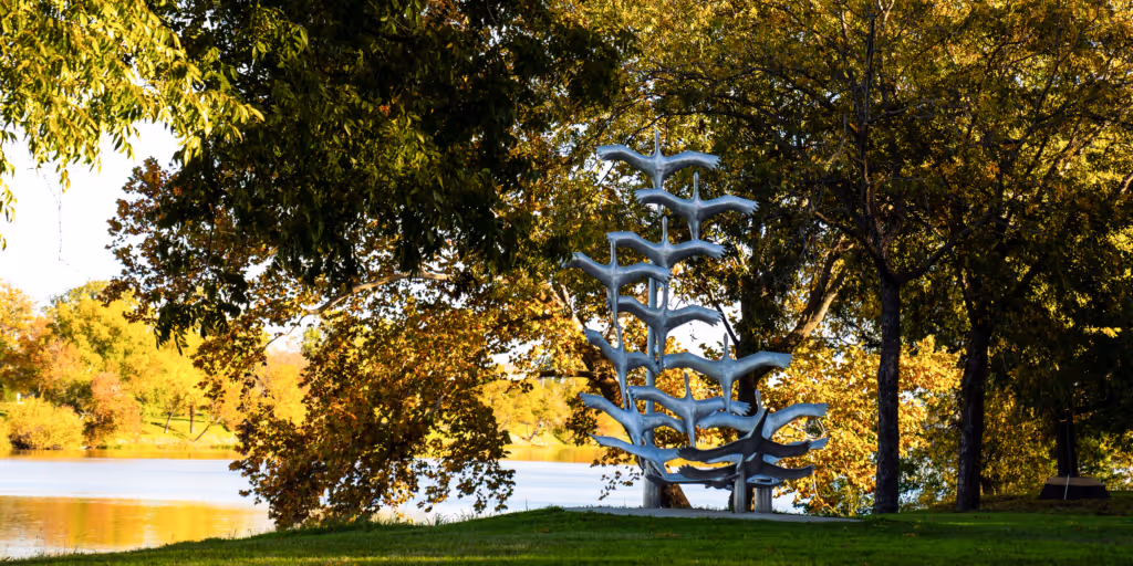Metal sculpture of birds in flight near a lake surrounded by trees with green and autumn foliage.