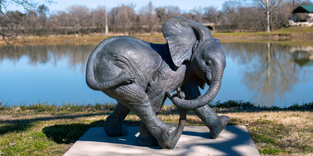 Gray sculpture of a walking baby elephant near a calm lake with trees in the background.
