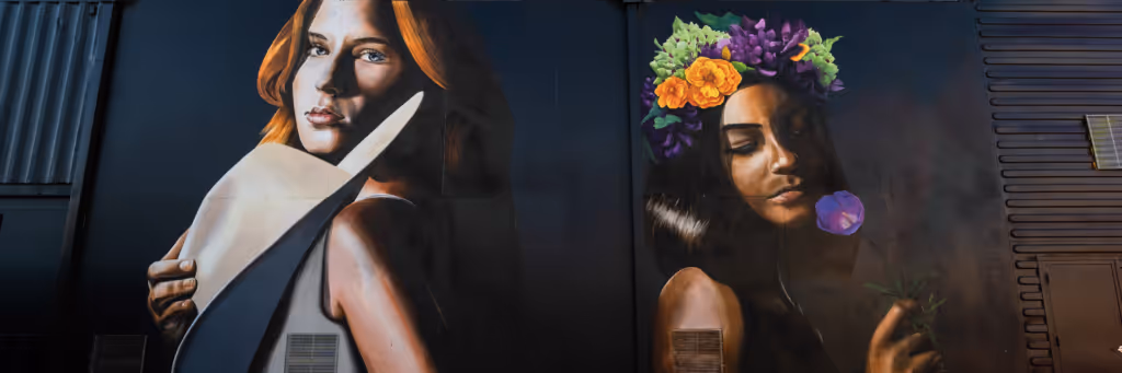 Street mural of two women: one with red hair holding a white guitar, and the other with a flower crown holding a purple flower.