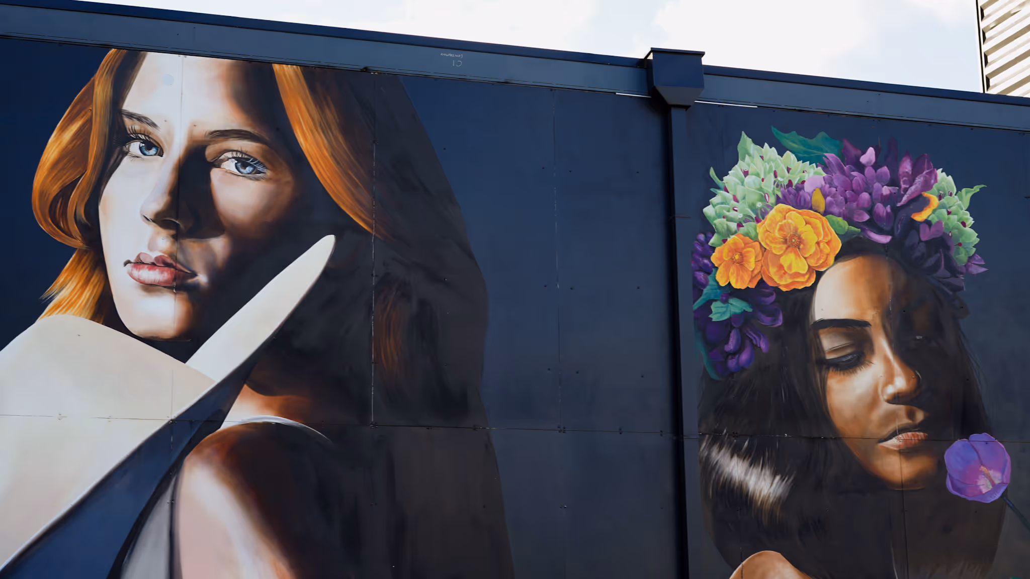 Mural on a dark wall showing two women: one with red hair and a serious expression, the other with dark hair wearing a colorful flower crown and holding a purple flower.