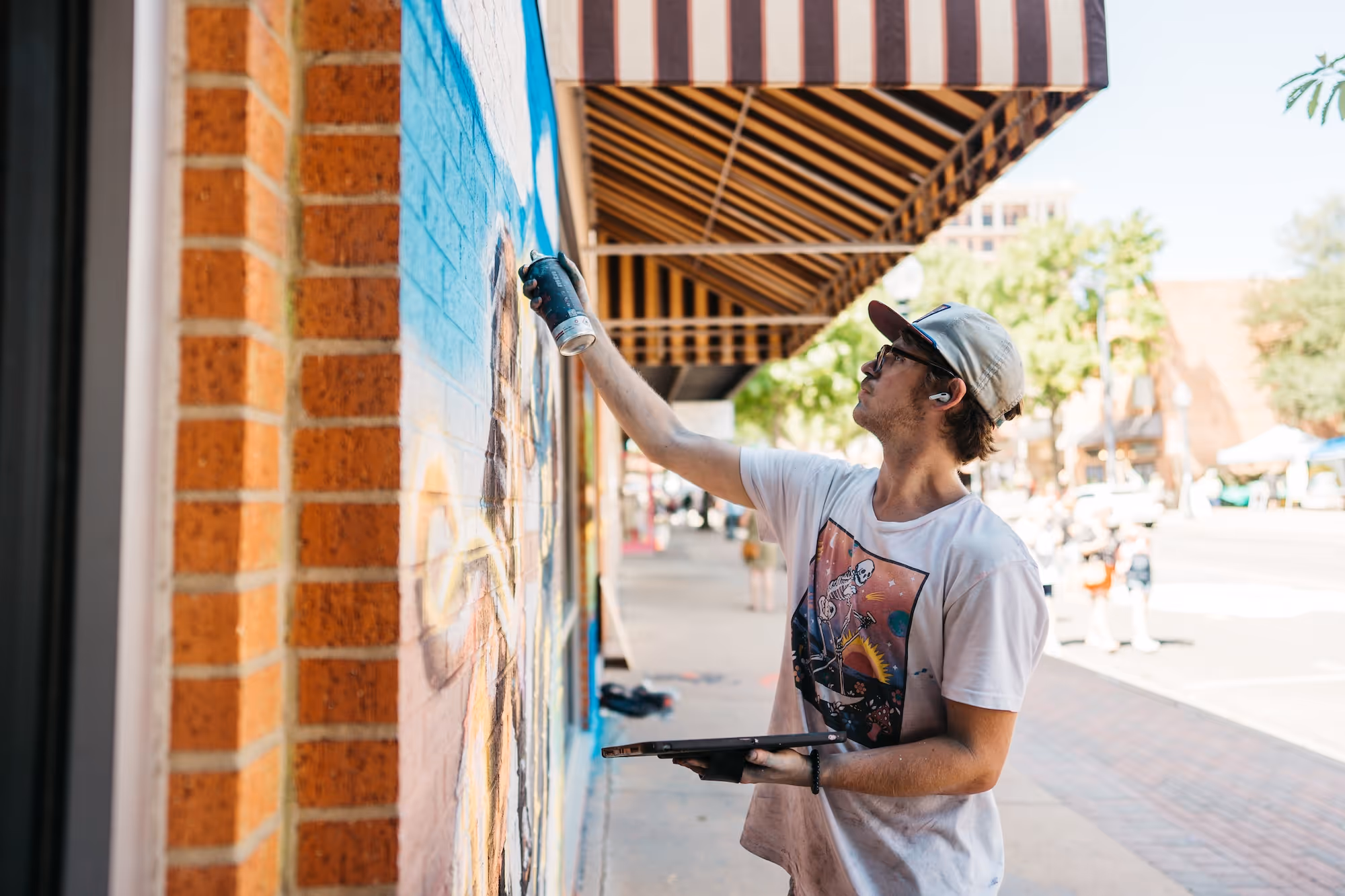 Young man wearing a backward cap and glasses spray painting a mural on a brick wall outdoors.
