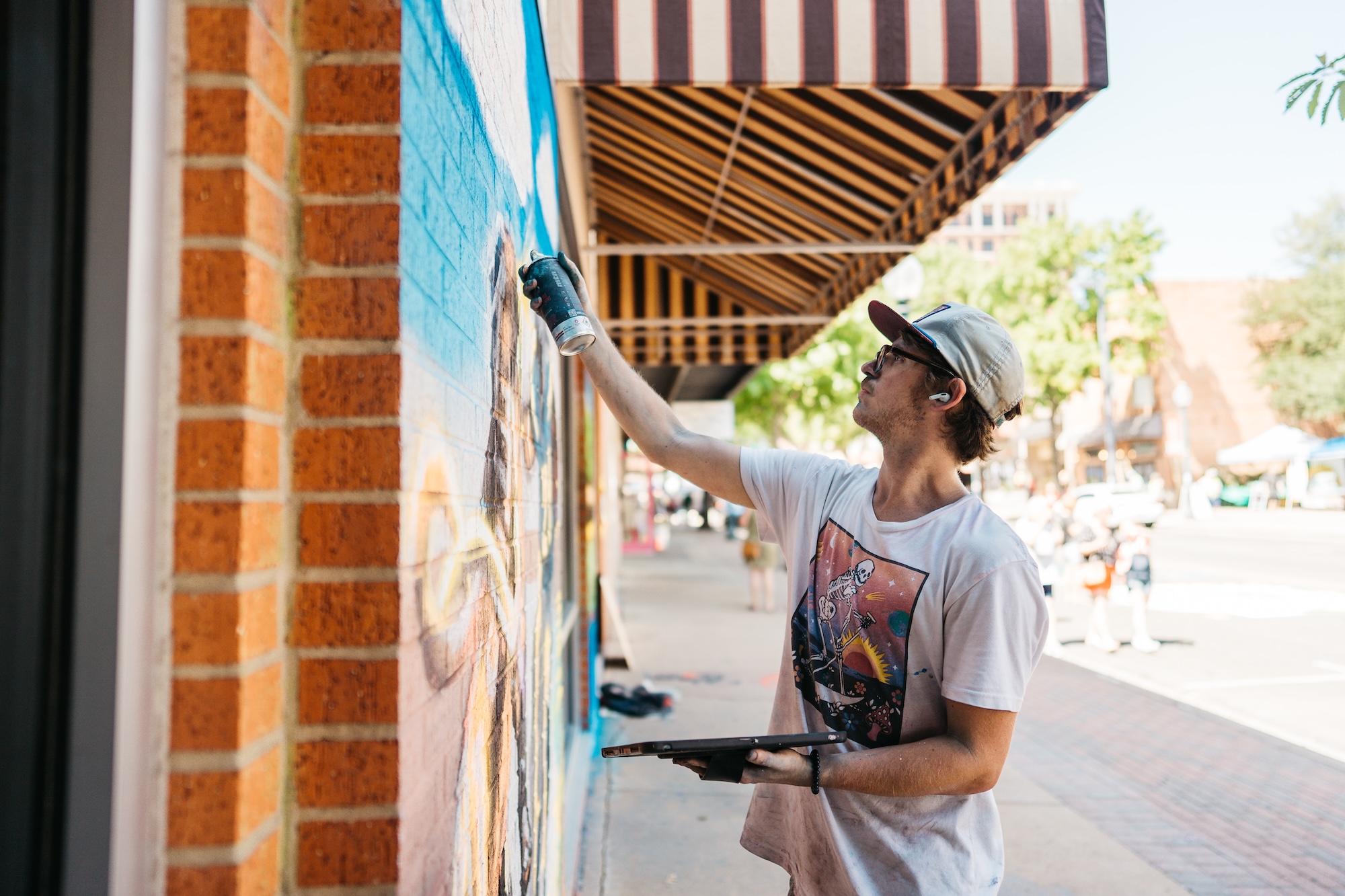 Young man wearing a backward cap and glasses spray painting a mural on a brick wall outdoors.